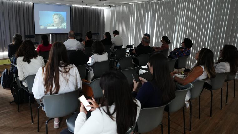 alt="Foto. Pessoas estão sentadas em cadeiras em uma sala escura com um telão."