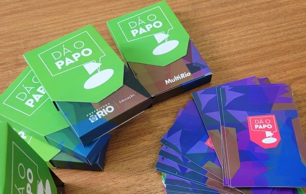 Foto. Caixas e cartas do jogo Dá o Papo estão dispostos sobre uma superfície de madeira. As caixas possuem om logotipo da MultiRio e da Prefeitura do Rio/ Educação.