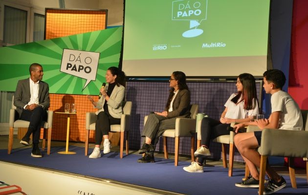 Foto. Cinco pessoas estão sentadas em cadeiras em um palco participando de uma discussão, sendo três adultos e dois estudantes de diferentes gêneros e raças. As cadeiras de cores claras estão dispostas em linha. No fundo, painéis coloridos, um deles com o logotipo do jogo Dá o papo, e um banner para projeção.