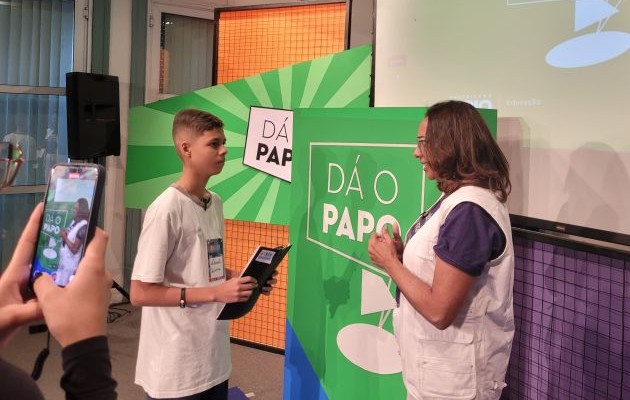 Foto. Um jovem estudante entrevista uma mulher adulta. No fundo, painéis coloridos, com o logotipo do jogo Dá o papo. Em primeiro plano, no canto esquerdo, uma pessoa segura um celular registrando a entrevista.